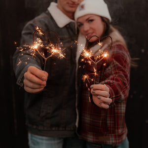 Close-up of a man and woman holding heart sparklers, their hands illuminated by the glowing gold sparks, against a beautifully blurred background.