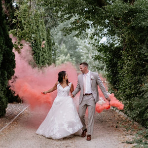 Man in tan suit and woman in wedding dress holding pink smoke bomb in each hand with large clouds of pink clouds trailing behind them