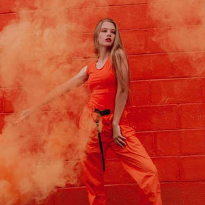 Photographer Smoke Bombs being used by woman wearing orange shirt and pants against orange wall holding an orange smoke bomb