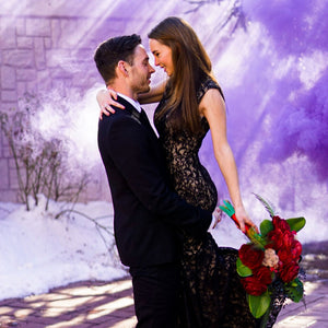 Man in black suit embracing woman in black wedding dress with purple color smoke bomb behind them creating smoky background