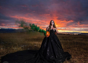 Lady wearing long black wedding dress holding carved pumpkin with green Halloween smoke bomb coming out eyes and nose for Halloween photo shoot