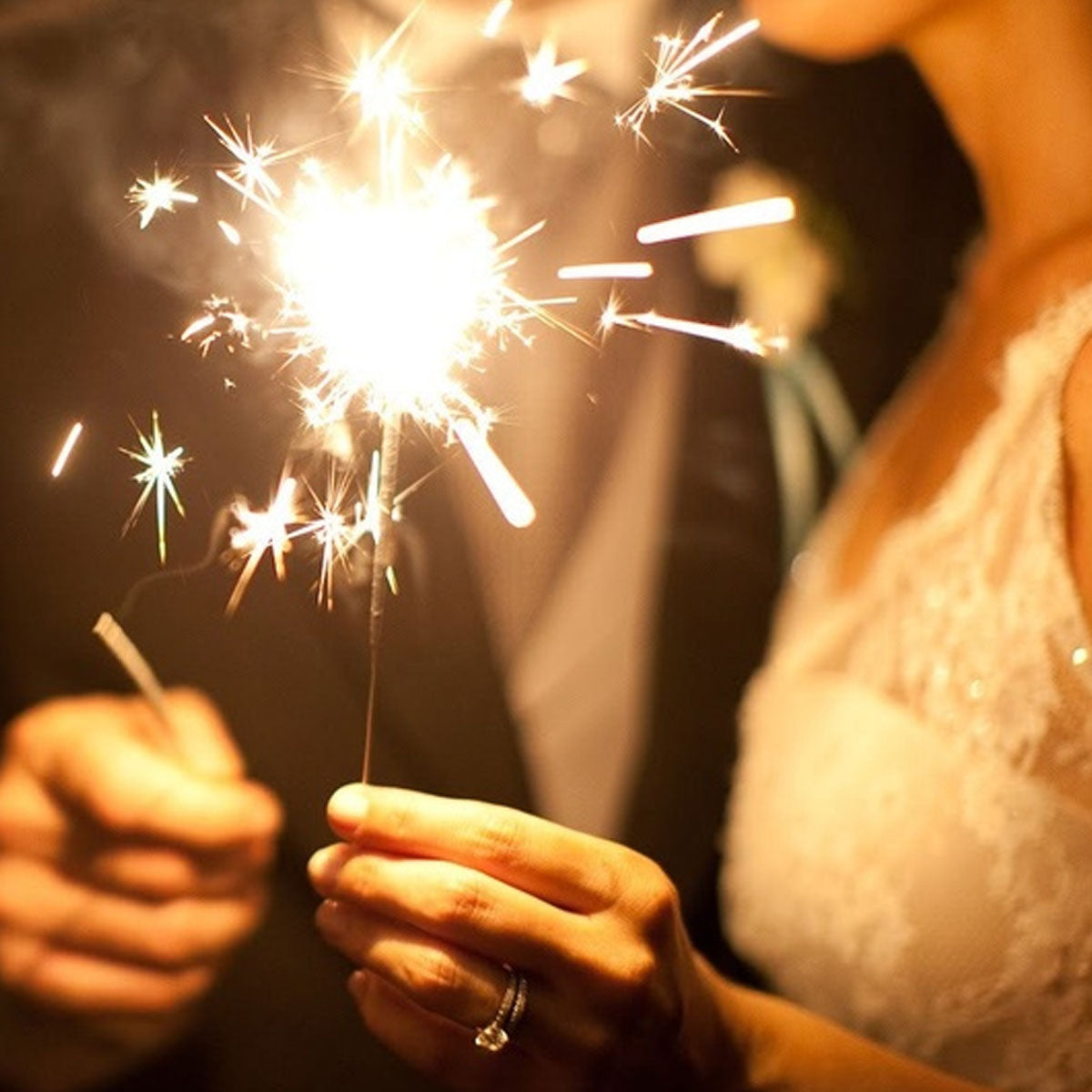 Close up of bride and groom holding 10-Inch Multi-Colored Sparklers emitting gold sparks against dark background