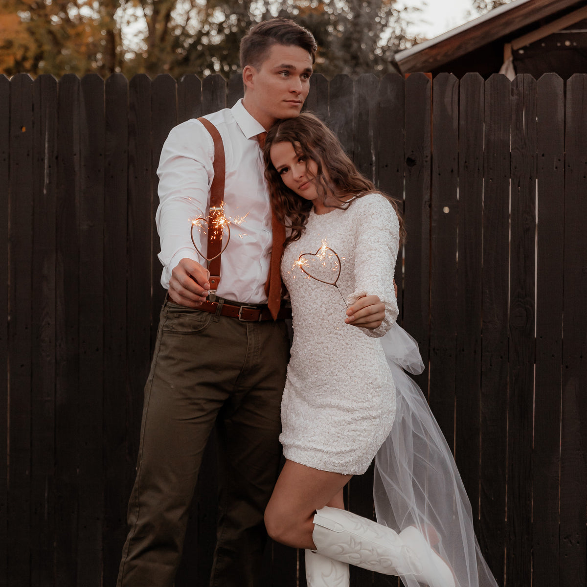 Guy dressed nice in slacks, white shirt and tie and lady wearing white formal dress holding heart sparklers in front of a brown fence