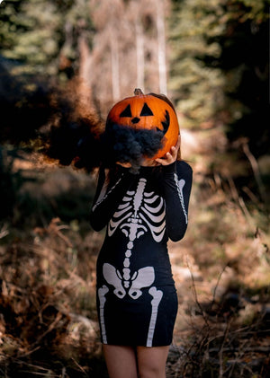 girl dressed in skeleton dress holding a pumpkin in front of her face with black smoke coming out it's eyes and mouth