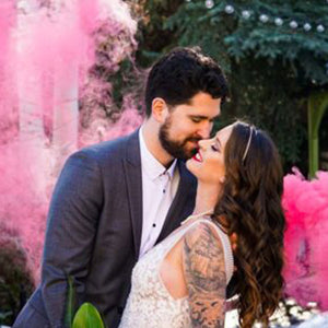Lady with tattoos  on arm smiling while man leans in for a kiss with pink smoke bomb creating clouds of pink background