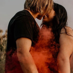 Man and woman in black shirts kissing while holding orange smoke bombs for photography 