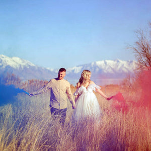 Husband and wife in open field with tall grass each holding pink and blue smoke bombs for gender reveal